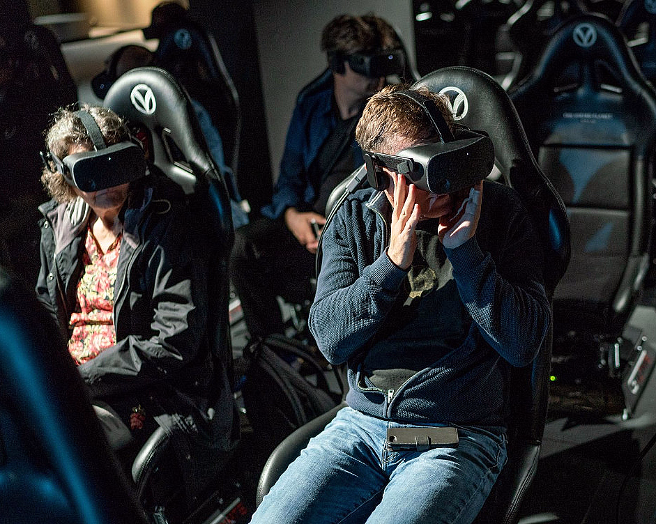 Personer med VR-briller i svarte stoler med V-logo på hodestøtten i et mørkt rom