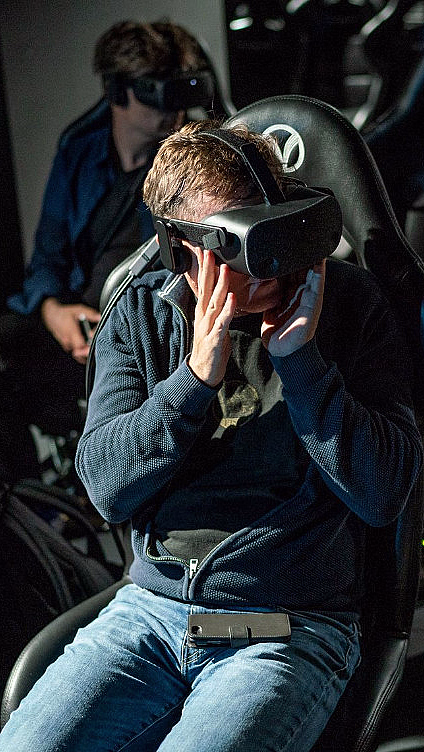 Personer med VR-briller i svarte stoler med V-logo på hodestøtten i et mørkt rom