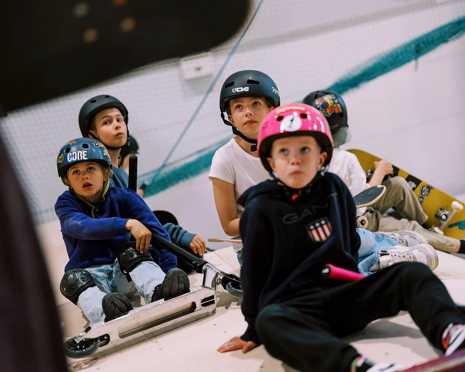 Barn med hjelmer sitter på rullebrett og sparkesykler i en skatepark, en hjelm merket CORE