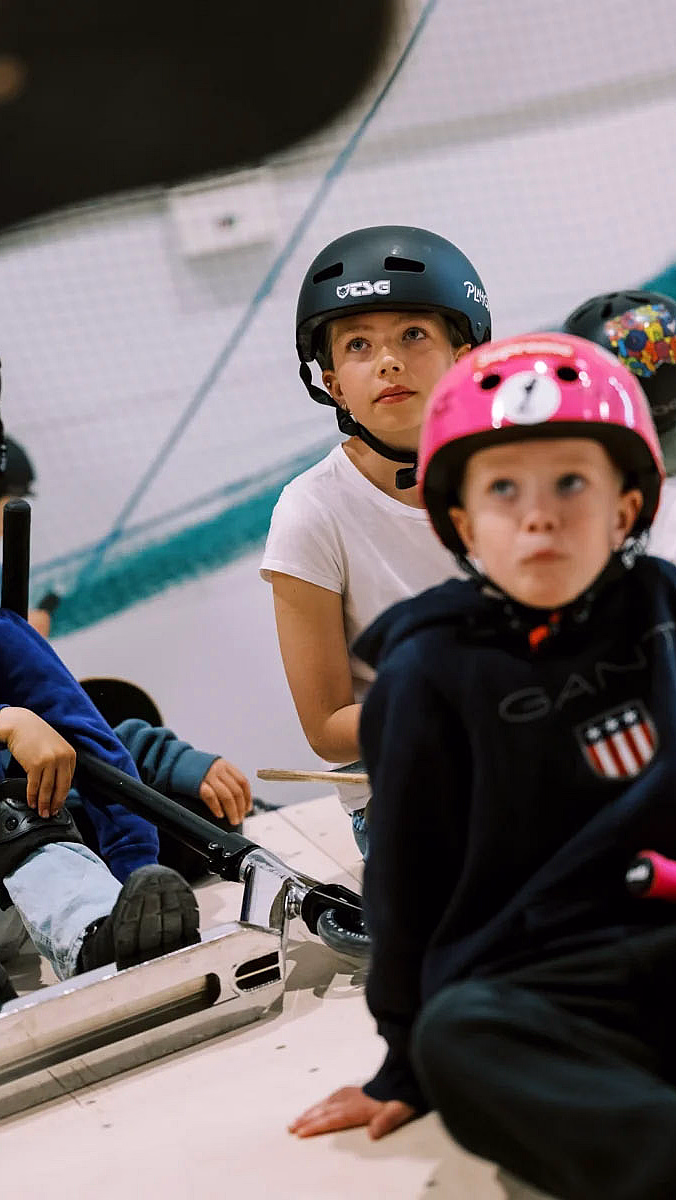 Barn med hjelmer sitter på rullebrett og sparkesykler i en skatepark, en hjelm merket CORE