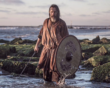 Person med sverd og rundt skjold i vannet ved mosegrodde steiner