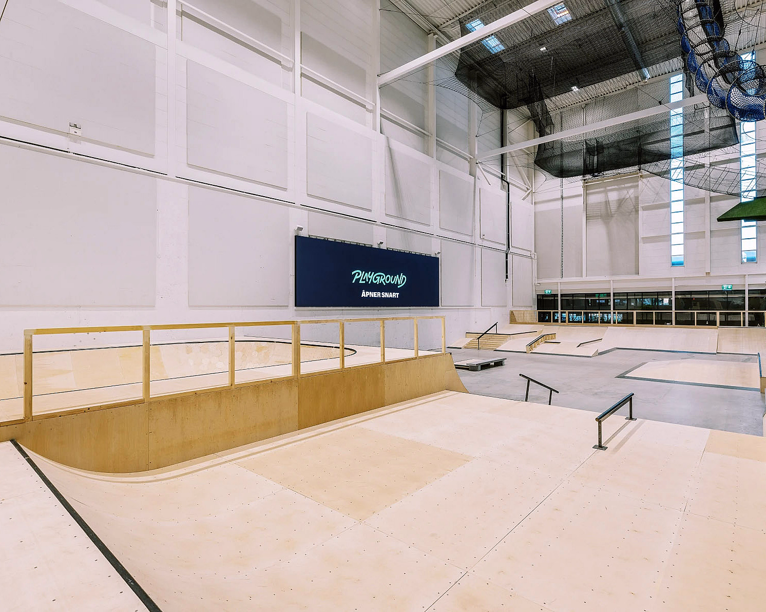 Innendørs skatepark med ramper i lyst tre og et blått skilt med teksten PLAYGROUND ÅPNER SNART