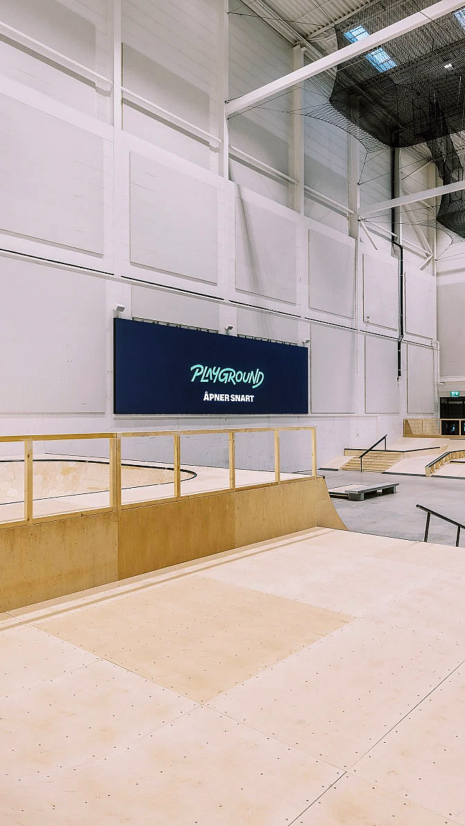 Innendørs skatepark med ramper i lyst tre og et blått skilt med teksten PLAYGROUND ÅPNER SNART