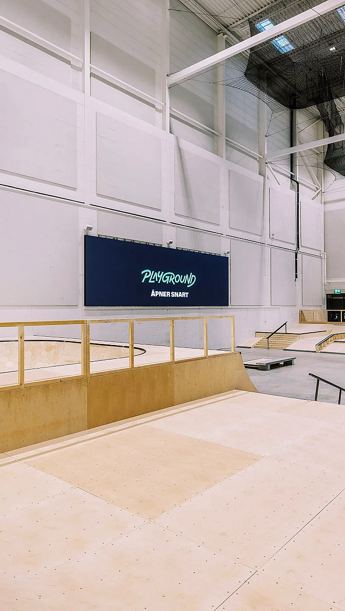 Innendørs skatepark med ramper i lyst tre og et blått skilt med teksten PLAYGROUND ÅPNER SNART