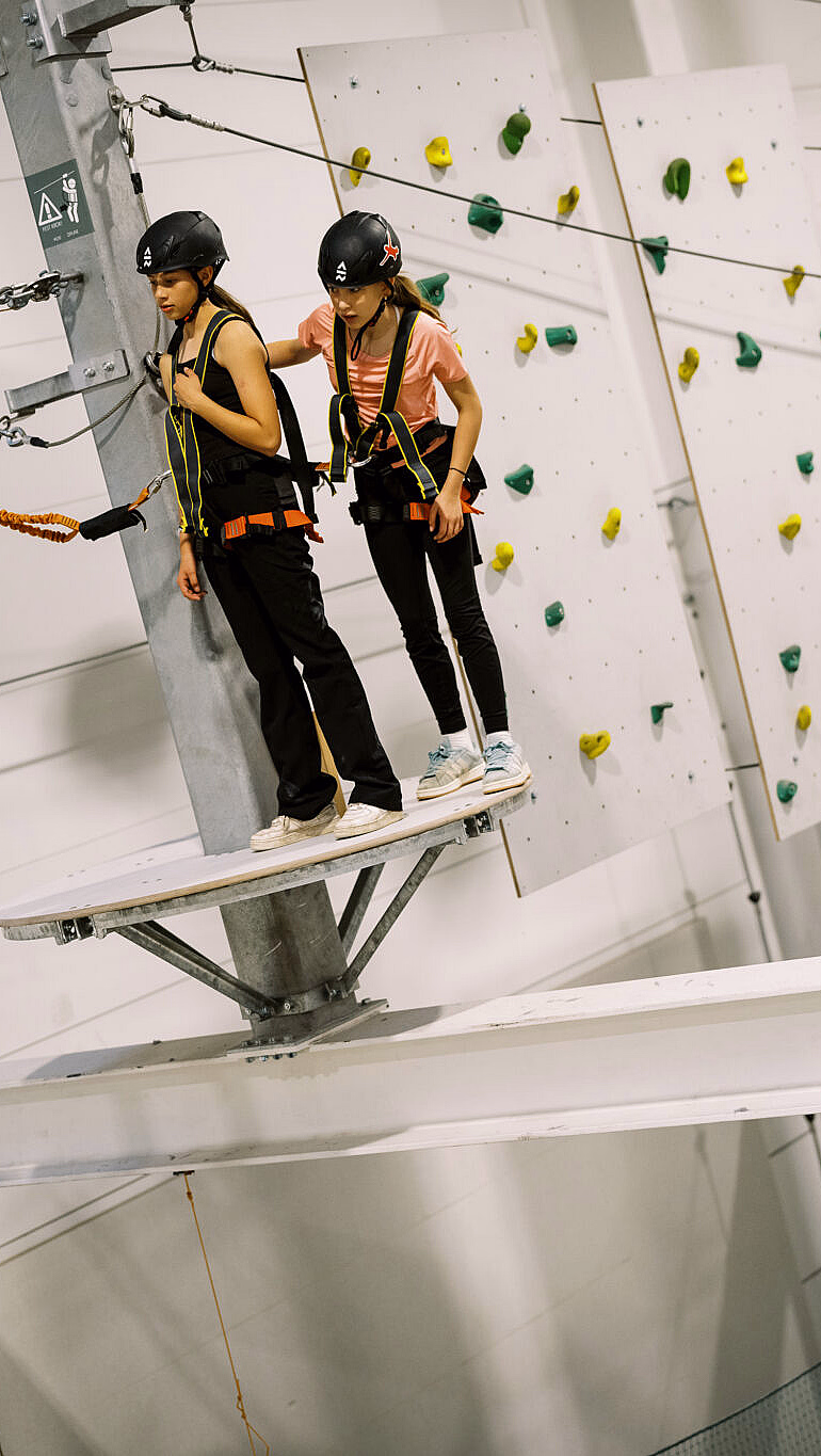 Personer med hjelm og klatresele på en innendørs klatrepark med zipline og klatrevegg