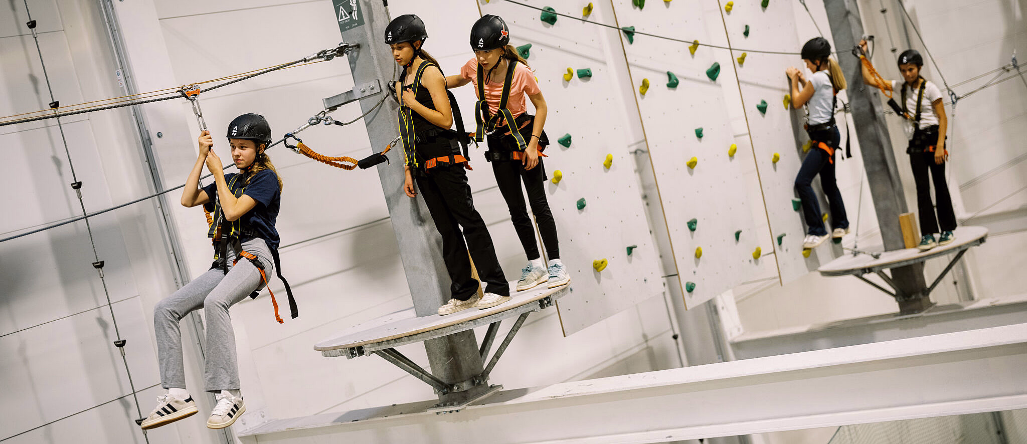 Personer med hjelm og klatresele på en innendørs klatrepark med zipline og klatrevegg