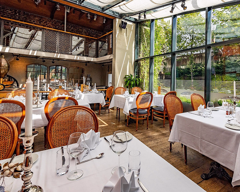 Lyst restaurantlokale med hvite duker og trestoler ved store vinduer med hageutsikt