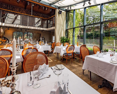 Lyst restaurantlokale med hvite duker og trestoler ved store vinduer med hageutsikt