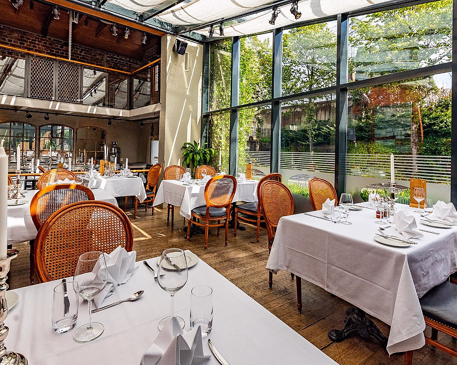 Lyst restaurantlokale med dekkede bord, trestoler og store vinduer mot grønne omgivelser