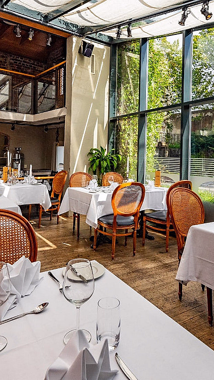 Lyst restaurantlokale med dekkede bord, trestoler og store vinduer mot grønne omgivelser