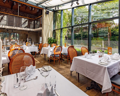 Lyst restaurantlokale med dekkede bord, trestoler og store vinduer mot grønne omgivelser