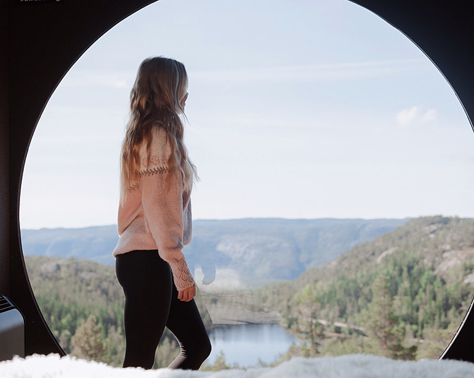 Person med langt hår og rosa genser ser ut av et stort rundt vindu mot skog og fjell