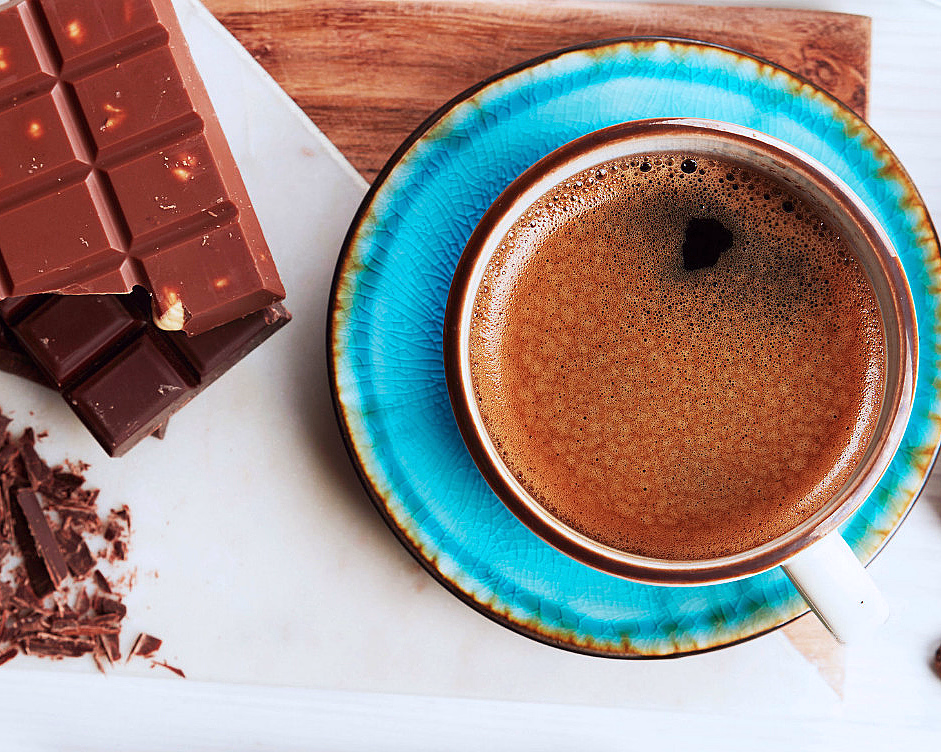 Kaffekopp på blått tefat ved siden av sjokoladeplater med nøtter og kakaobønner