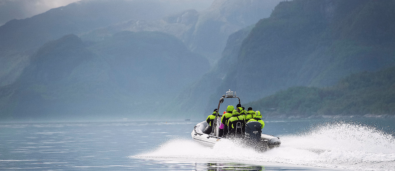 Hurtiggående motorbåt med personer i grønne jakker foran tåkete fjell