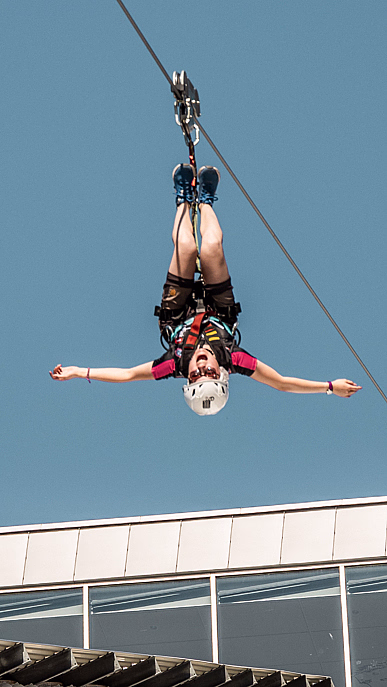 Person i rød skjorte henger opp ned på en zipline med utstrakte armer mot en blå himmel