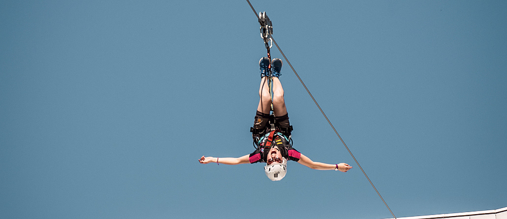 Person i rød skjorte henger opp ned på en zipline med utstrakte armer mot en blå himmel