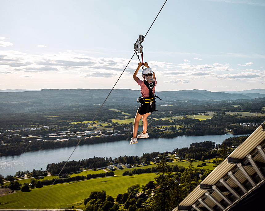 Person i sele og hjelm som kjører zipline over en elv og grønt landskap