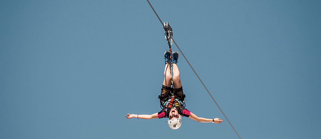 Person som henger opp ned på en zipline med utstrakte armer mot en blå himmel