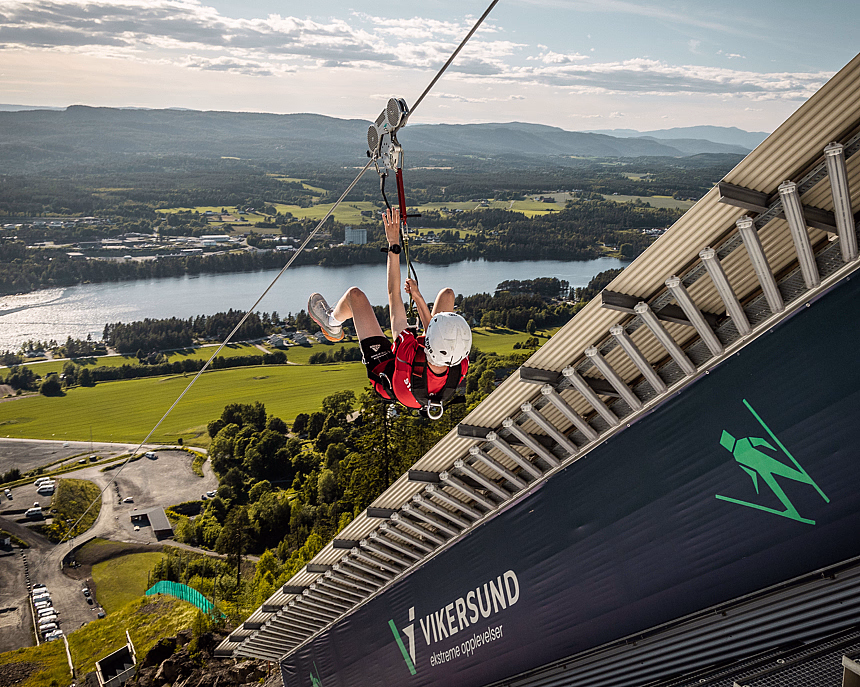 Person i rød skjorte og hjelm på zipline fra tårn merket Vikersund over grønt landskap og innsjø