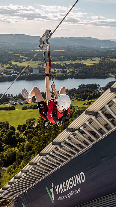 Person i rød skjorte og hjelm på zipline fra tårn merket Vikersund over grønt landskap og innsjø