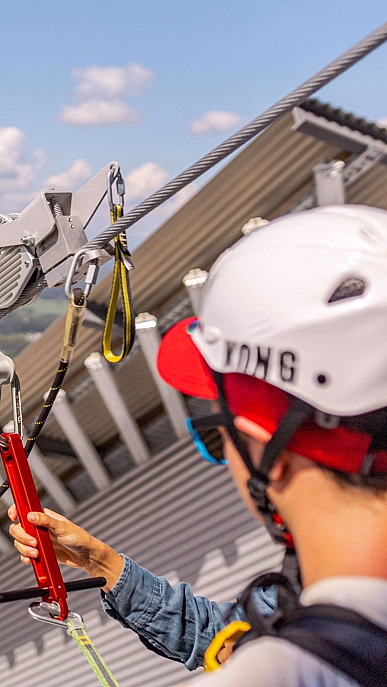 Person med hvit hjelm og rød caps holder et rødt håndtak på en zipline over en innsjø