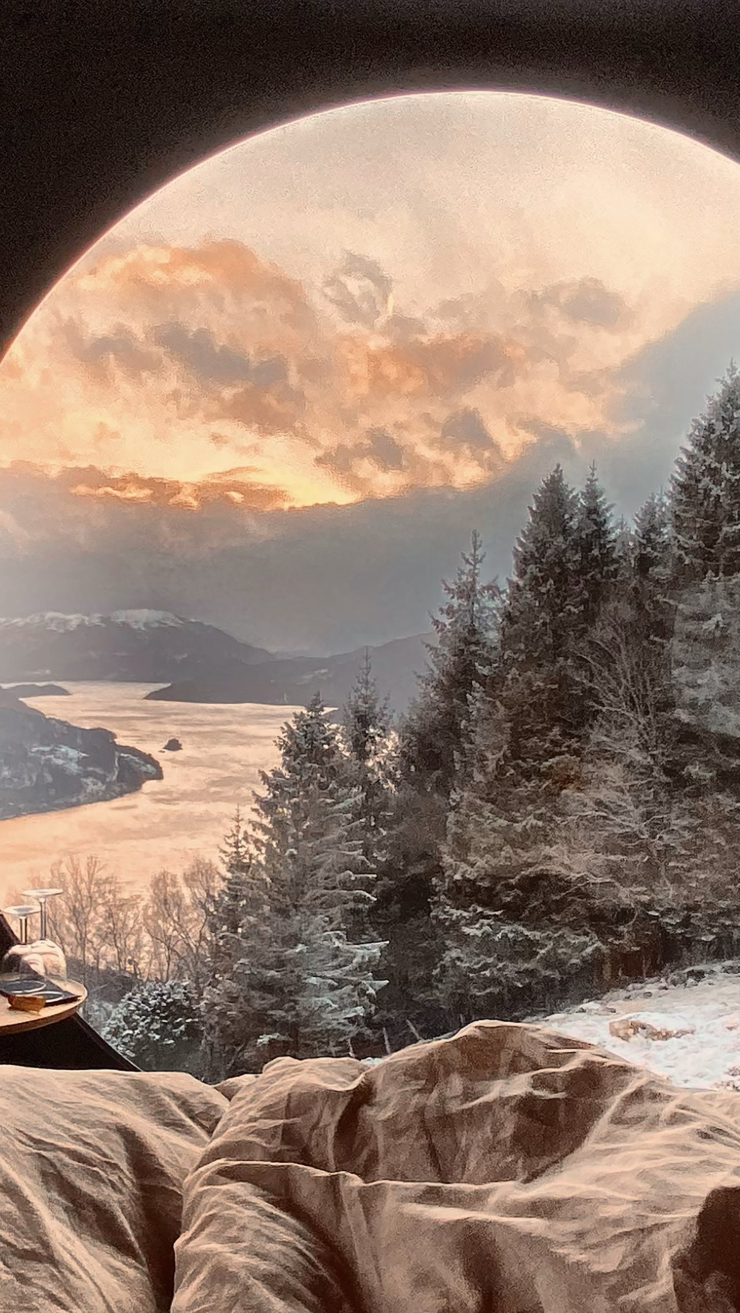 Seng ved et rundt vindu med utsikt til snødekte fjell i solnedgang