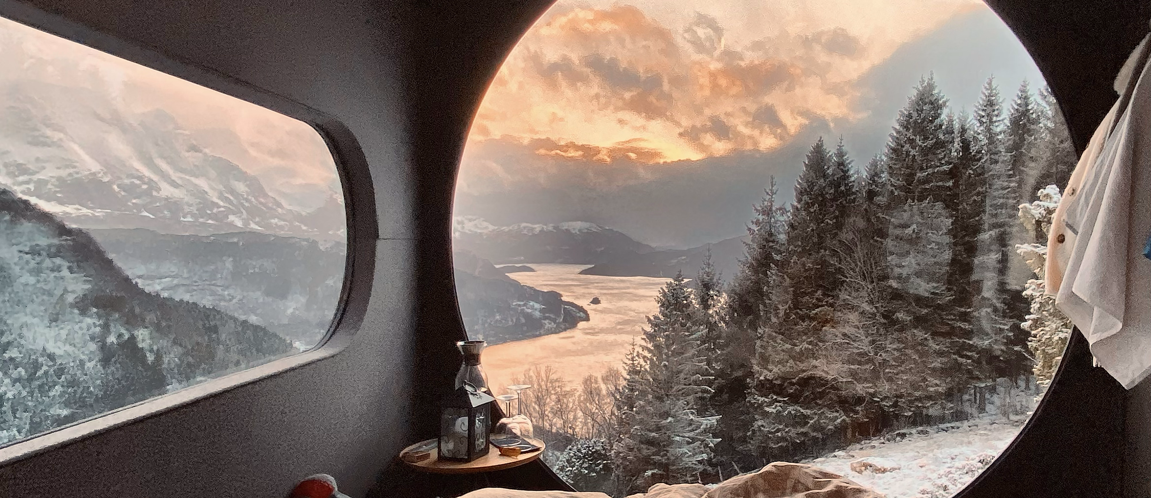 Seng ved et rundt vindu med utsikt til snødekte fjell i solnedgang