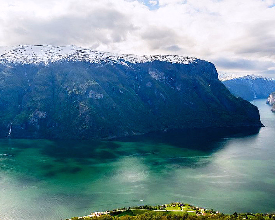 Snødekte fjell over en grønn fjord med en foss og små hus ved vannet