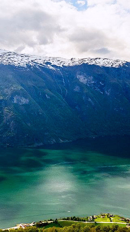Snødekte fjell over en grønn fjord med en foss og små hus ved vannet
