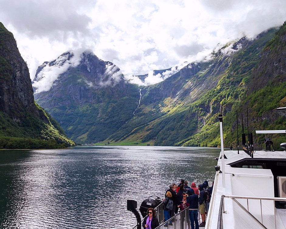 Passasjerer på en båt i en fjord omgitt av bratte fjell med tåke og en foss