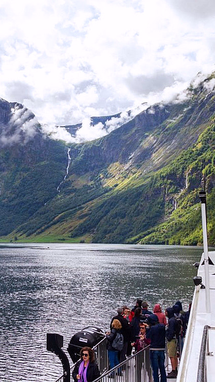 Passasjerer på en båt i en fjord omgitt av bratte fjell med tåke og en foss