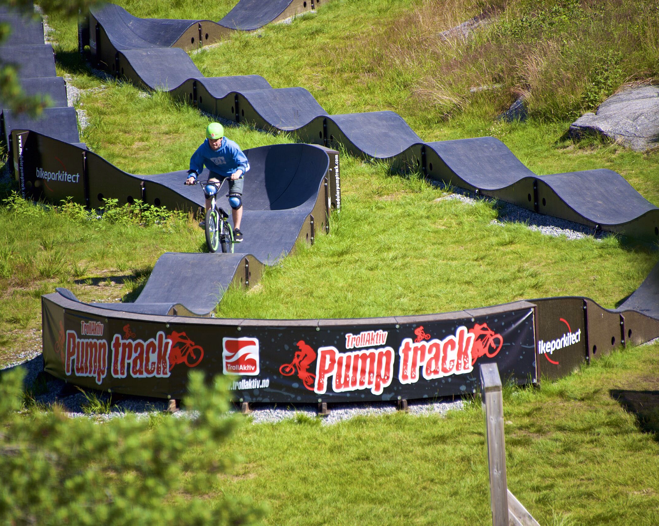 Syklist i blå skjorte og grønn hjelm på en bølgete pumptrack med banneret Pump track