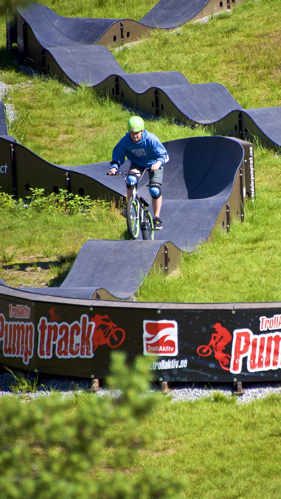 Syklist i blå skjorte og grønn hjelm på en bølgete pumptrack med banneret Pump track
