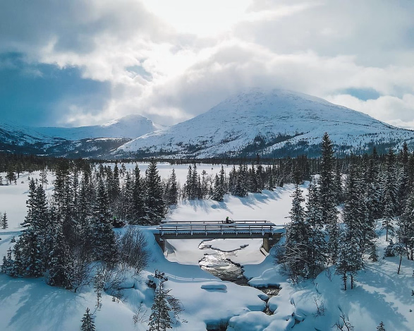 Snøscooter på en bro i et snødekt fjellandskap med granskog