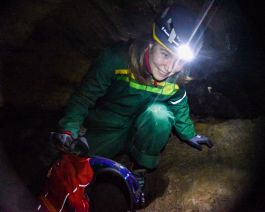 Kvinne i grønn kjeledress og hjelm med tent lykt inne i en mørk grotte