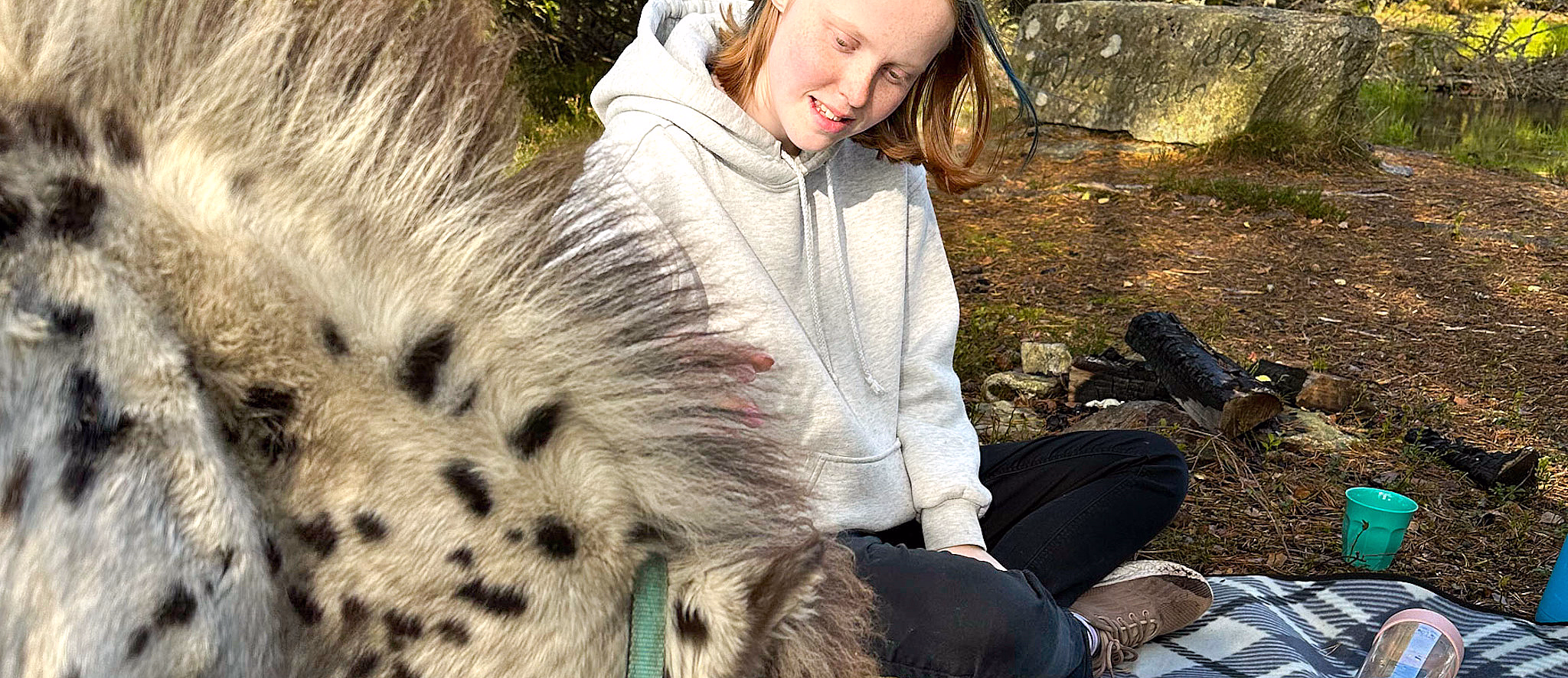 Ung person på rutete teppe i skogen med et lite prikkete dyr som spiser fra en papirpose
