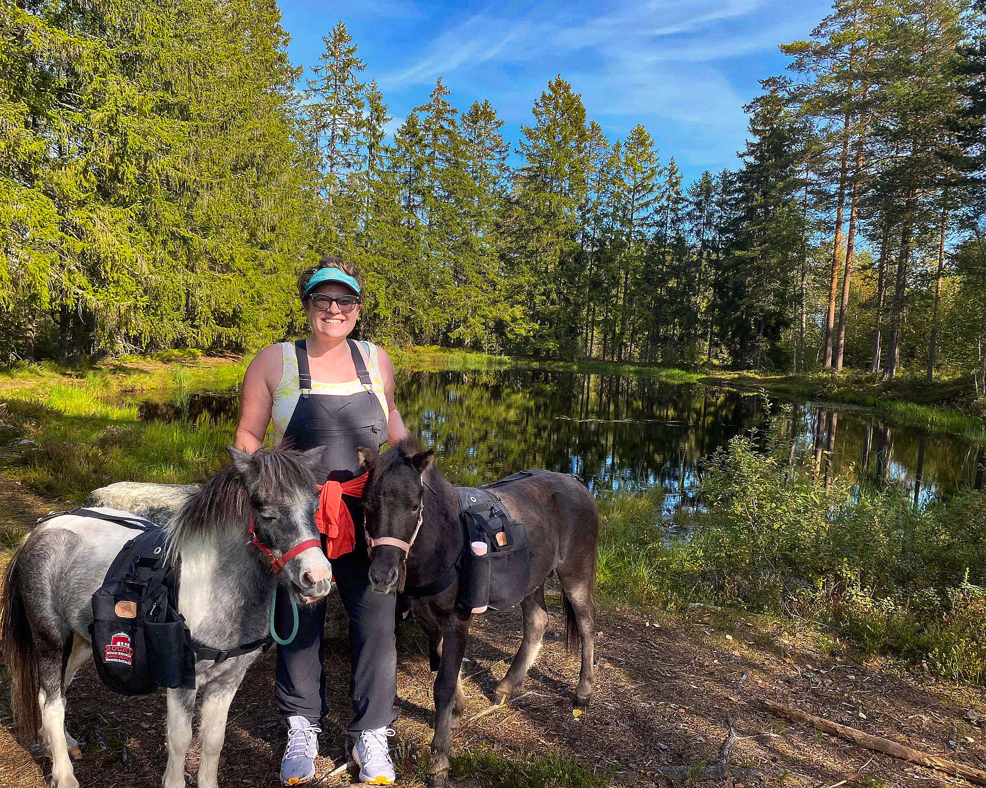Smilende person med en grå og en svart ponni med salvesker ved et vann i skogen