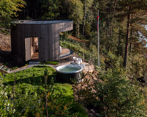 Rund trehytte i skogen ved en innsjø med badestamp og en blå kajakk ved en brygge