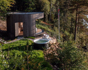 Rund trehytte i skogen ved en innsjø med badestamp og en blå kajakk ved en brygge