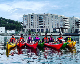 Gruppe i fargerike kajakker på vannet foran moderne leilighetsbygg