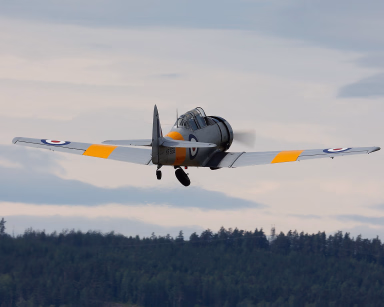 Grått og gult propellfly sett bakfra under takeoff over skogkledde åser