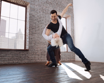 En mann og en kvinne danser tango i et rom med murvegg og store vinduer