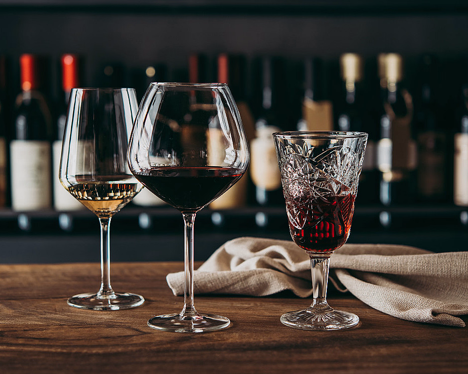 Tre ulike glass med vin på et trebord foran en hylle med vinflasker