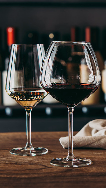 Tre ulike glass med vin på et trebord foran en hylle med vinflasker