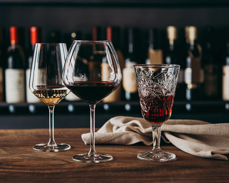 Tre ulike glass med vin på et trebord foran en hylle med vinflasker