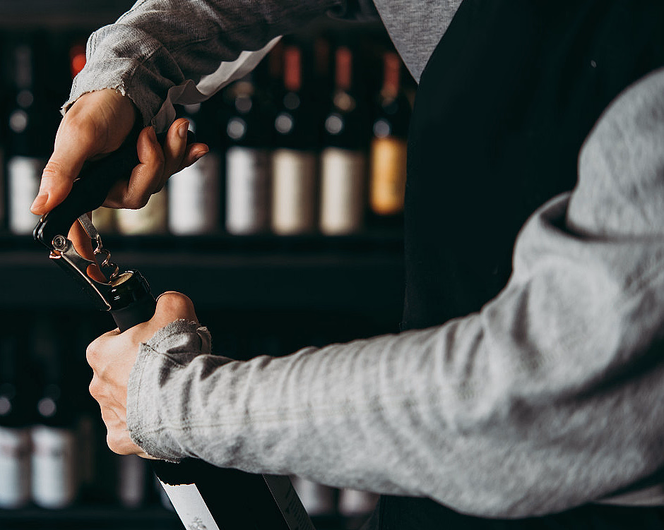 En person som åpner en vinflaske med en korketrekker foran en hylle med vinflasker