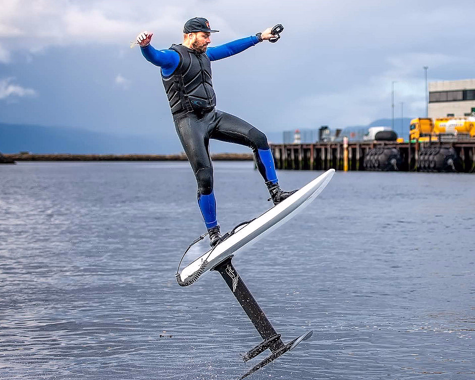 Person i våtdrakt på et hydrofoil-surfebrett over vannet med en brygge i bakgrunnen