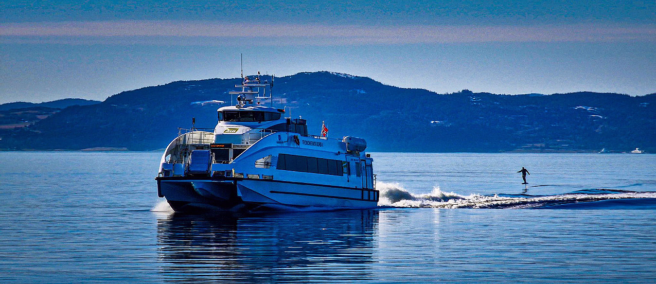 Hvit ferge på vannet og en person på hydrofoil i kjølvannet med fjell i bakgrunnen