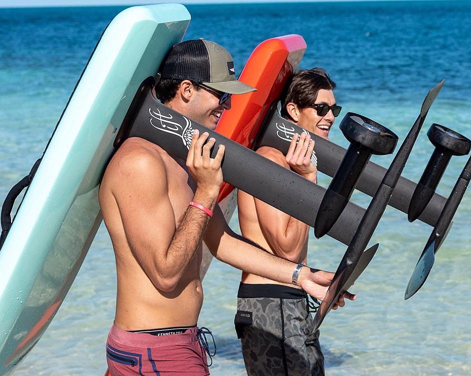 To menn bærer et rødt og et lyseblått hydrofoil-surfebrett på en strand