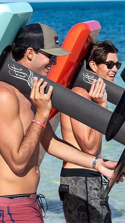 To menn bærer et rødt og et lyseblått hydrofoil-surfebrett på en strand